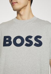Camiseta de algodón gris con gran texto bordado en azul marino que dice "BOSS". Mangas cortas y escote redondo. Textura suave.