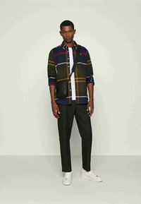 Bărbat îmbrăcat cu o cămașă în carouri multicolore, peste un tricou alb, pantaloni negri, adidași albi și o geantă crossbody neagră, stând în fața unui fundal simplu.