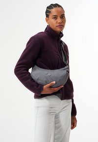 Graue, leichte Schultertasche mit runder Form, aus strukturiertem Stoff und zwei runden Knöpfen. Quer über den Körper getragen mit einem schwarzen Riemen.