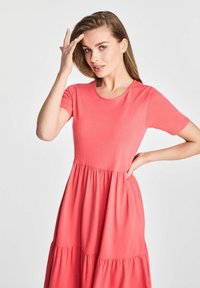 Koraliowy różowa sukienka z okrągłym dekoltem, krótkimi rękawami i warstwową, rozkloszowaną spódnicą. Wykonana z miękkiego, lekkiego materiału o gładkiej teksturze.