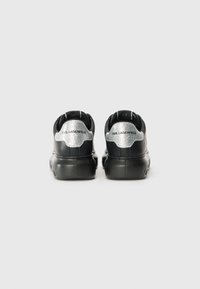 Black sneakers featuring a glossy texture, silver metallic accents on the heel, and a logo stamped on the back. Rounded design with thick sole.