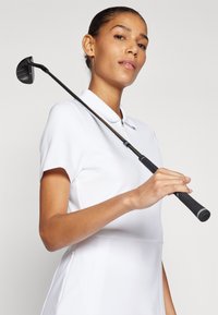 Golf klub z črno glavo in oprijemom, ki ga drži oseba v beli polo srajci s kratkimi rokavi in z zadrgo spredaj. Gladka tkanina, čiste linije.
