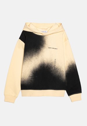 Beiger Kapuzenpullover mit schwarzem Spray-Paint-Muster, Schriftzug "Calvin Klein Jeans" auf der Brust und im unteren Vorderbereich, lange Ärmel, gerippte Bündchen und Saum.
