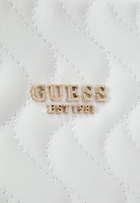 Bílý prošívaný materiál s jemným prošíváním, uprostřed výrazně zobrazené zlaté kovové logo s nápisem "GUESS EST 1981".