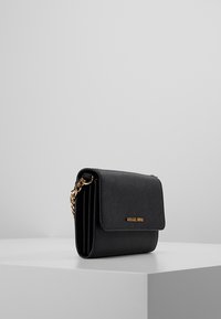 Bolso cruzado de piel negra con cierre de solapa, correa de cadena dorada y un acento de logo en la parte frontal. Superficie texturizada y diseño compacto.