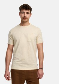 Camiseta de manga corta color crema con un bolsillo en el pecho, cuello redondo y textura sutil. Acentuada con un pequeño logo cerca del bolsillo.