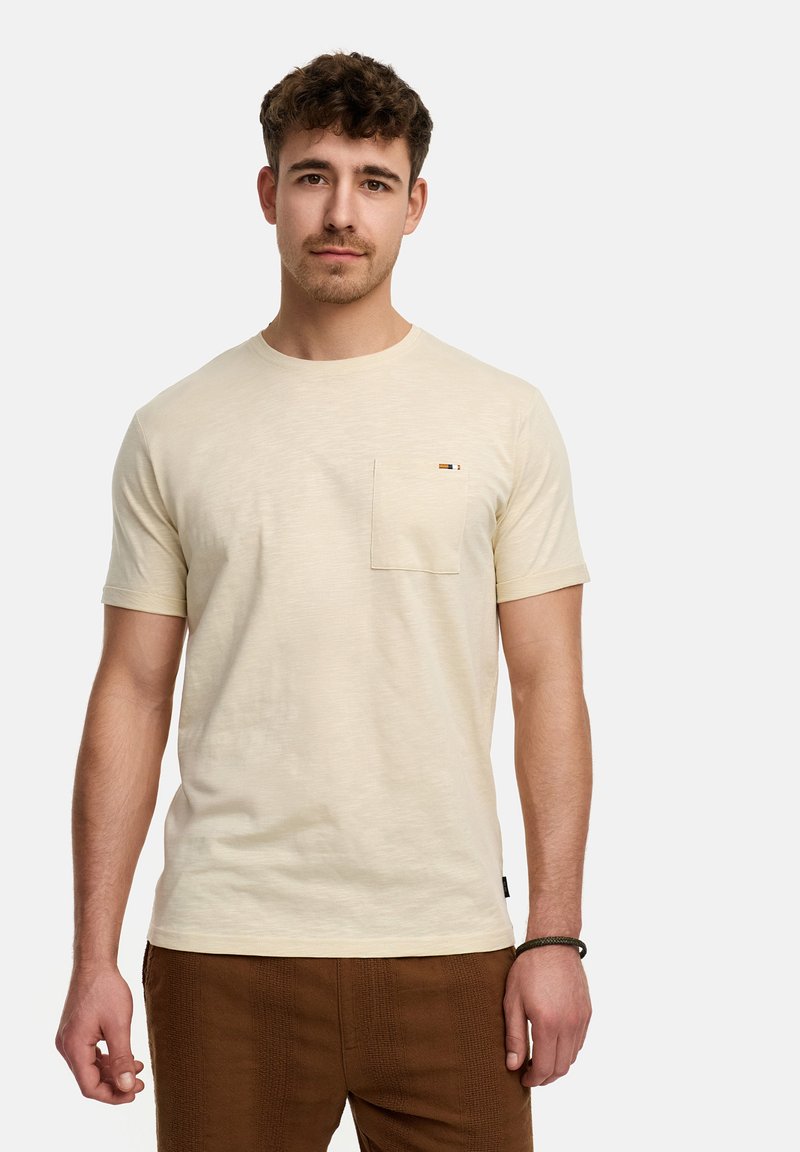 Camiseta de manga corta color crema con un bolsillo en el pecho, cuello redondo y textura sutil. Acentuada con un pequeño logo cerca del bolsillo.