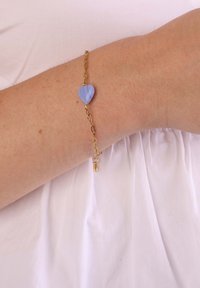Bracelet en chaîne dorée avec un perle en forme de cœur bleu clair, et un petit détail de verrou près du fermoir.