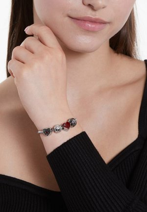 Silberne Charm-Armband mit Herz- und Rosen-Anhängern, akzentuiert mit rotem Emaille und einem klaren Kristall. Wird an einer Hand mit einer weichen, glatten Textur getragen.