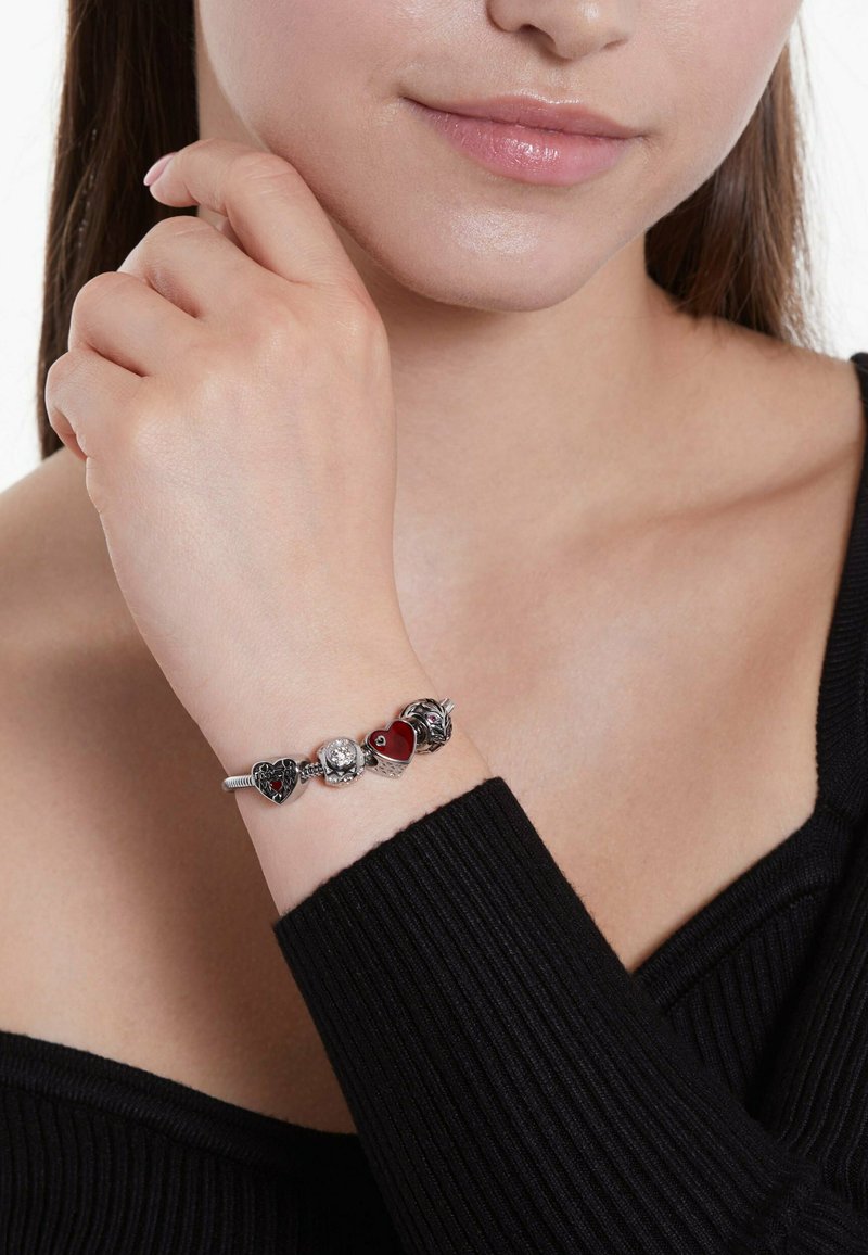 Zilveren bedelarmband met hart- en rozenbedels, aangevuld met rode emaille en een heldere kristal. Draag het aan een hand met een zachte, gladde textuur.