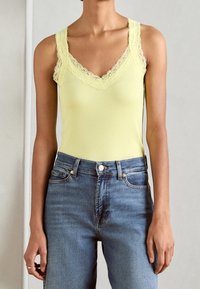 Kvinna i en ljusgul linne med spetsdetaljer, nedstoppad i högmidjade blå jeans, som står framför en neutral bakgrund.