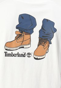 White t-shirt featuring an illustration of two brown Timberland boots with laces and blue jeans. "Timberland" logo below the design.