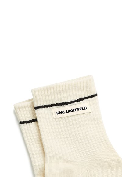 Chaussettes côtelées couleur crème avec une bande noire près du haut et une étiquette rectangulaire blanche portant la mention « KARL LAGERFELD » en texte noir.