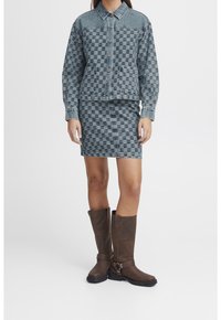 Kobieta ubrana w niebieską kratkowaną dżinsową kurtkę i dopasowaną spódnicę, z brązowymi skórzanymi kozakami za kolano, stojąca na białym tle.