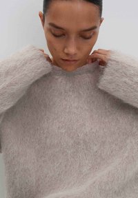 Woman with dark hair adjusts the neckline of her soft, fluffy light gray sweater against a plain gray background.