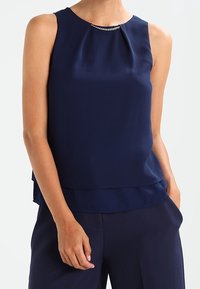 Mörkblå ärmlös topp i mjukt tyg med rund halsringning och en diskret veckdetalj samt en dekorativ metallackcent.