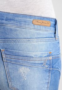 Světle modré džíny z denimu se vybledlým povrchem, s viditelnými švy, koženou etiketou a jemným opotřebením v blízkosti kapsy.