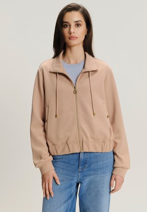 Svijetloružičasta jakna na zip s visokim ovratnikom, detaljem na vezanje i zlatnim akcentima. Rebraste manžete i rub dodatno naglašavaju njen prilagođeni izgled.