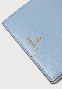 Gaiši zila ādas maku ar teksturētu apdari, ar zelta "FURLA" logo un melnu šuvumu gar maliņām.