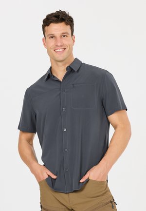 Glurende man die een donkergrijs overhemd met korte mouwen en beige cargobroek draagt, met zijn handen in de zakken.