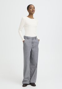 Sieviete stāv ar rokām kabatās, ģērbusies baltā garā piedurkņu blūzē, platās pelēkās džinsās un melnās kurpēs uz baltas fona.