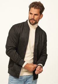 Schwarze Wildleder-Bomberjacke mit gerippten Bündchen und Kragen, Reißverschluss, Seitentaschen, getragen über einem hellbeigen Rollkragenpullover.