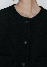 Mustat teksturoidut neuleet, joissa on pyöreä kaula-aukko, neljä etupainiketta ja kaksi tikkutaskua. Kangas on löyhärakenteinen.