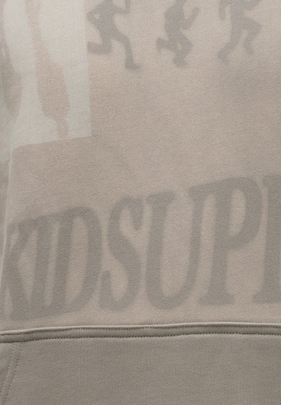 Ύφασμα φούτερ σε μπεζ χρώμα με ξεθωριασμένα γραφικά και κείμενο, που περιλαμβάνουν το "KIDSUP", με υφή και ορατές ραφές.