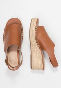 Sandales à talons compensés en cuir marron avec une semelle en jute tissée. Design à bout ouvert, bride à l'arrière avec un accent métallique et détails de couture.