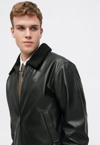 Mustmust mustrindast tekstuuriga musta lambanaha krae, lukuga ette sulgumine ja küljetaskud. Omab rinnanupul peenelt logo.