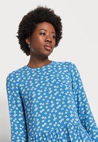 Vestido azul de manga longa com padrão floral branco, decote redondo e saia franzida. Feito de um tecido leve com textura suave.