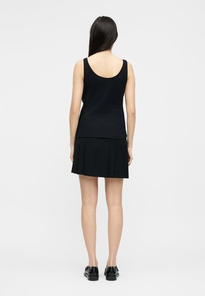 Γυναίκα με μακριά σκούρα μαλλιά, φορώντας μαύρο αμάνικο τοπ, μαύρη πλισέ φούστα και μαύρα γυαλιστερά παπούτσια, στέκεται με την πλάτη προς τα εμπρός σε απλό φόντο.