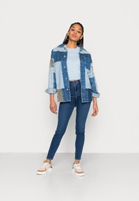 Denimowa koszula overshirt w odcieniach niebieskiego, noszona na jasnoniebieskim swetrze, w połączeniu z ciemnymi dżinsami skinny oraz trampkami z beżowym akcentem.