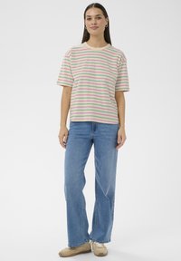 Frau trägt ein cremefarben gestreiftes T-Shirt, blaue Jeans und beige Turnschuhe, steht vor einem schlichten weißen Hintergrund.