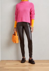 Pink ribbed sweater over an orange turtleneck, black leather leggings, black loafers, and an orange handbag, standing on wooden floor.