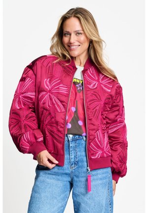 Vrouw glimlacht terwijl ze een roze bomberjack draagt met grote bloemenborduursels over een grafisch T-shirt en blauwe spijkerbroek tegen een witte achtergrond.