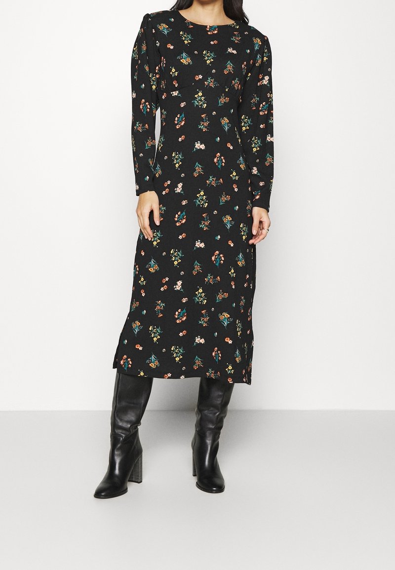 Femme portant une robe midi noire à manches longues avec un imprimé floral coloré et des bottes noires à talons montant jusqu'au genou, debout devant un fond blanc.