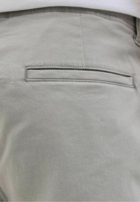 Gros plan sur un pantalon gris clair montrant une seule poche passepoilée à l'arrière et une partie d'une ceinture blanche au-dessus.