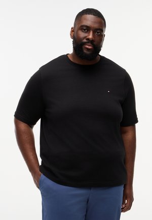 Mann med skjegg iført svart kortermet skjorte med liten logo og blå bukser, stående med en hånd i lommen mot hvit bakgrunn.