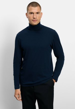 Man met kort haar die een donkerblauwe coltrui en zwarte broek draagt, staand tegen een effen witte achtergrond.
