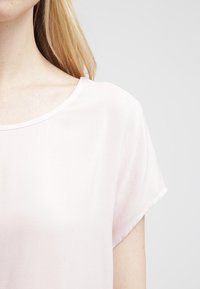 Světle růžová košile s krátkým rukávem a kulatým výstřihem, hladká textura a uvolněný střih. Minimalistický design bez viditelných vzorů či ozdob.