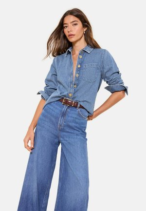 Naine kannab nööpidega denimshirt'i, millel on kuldsed nupud, ja kõrge vöökohaga laia säärega teksapükse pruuniga vööga, poseerides ühe käega puusal.