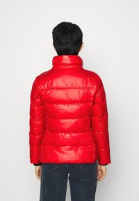 Rød puffjakke med en skinnende finish, horisontalt quiltet design, høj krave og en lavere bageste kant. Båret over mørke jeans.