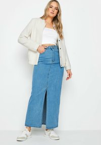 Giacca leggera crema sopra un crop top bianco, abbinata a una maxi gonna di jeans a vita alta con spacco frontale e sneakers bianche.
