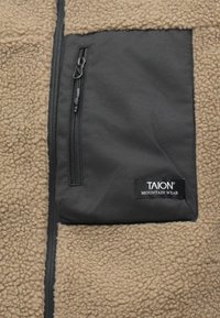 Κρεμ φλις μπουφάν με μαύρη τσέπη με φερμουάρ που φέρει την ετικέτα "TAION MOUNTAIN WEAR". Μαλακή υφή με αντίθετες περιοχές υφάσματος.