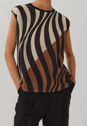 Mouwloze top met zwarte, beige en bruine golvende verticale strepen, gedragen met een zwarte broek, handen in de zakken tegen een effen achtergrond.