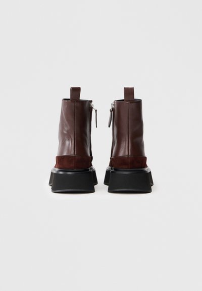 Een paar donkerbruin leren enkelboots met suède accenten, zilveren zijritsen, trektabs en dikke zwarte platte zolen, gezien vanaf de achterkant.