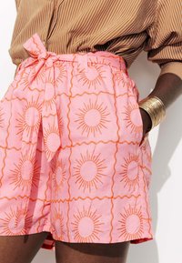 Shorts roses avec un motif de soleil en broderie orange, dotés d'une taille à nouer et de poches latérales, fabriqués en tissu léger.