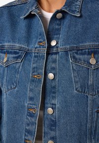 Denim jaka vidējā zilā krāsā ar sudraba pogām, krūšu kabatām un dzeltenām šuvēm. Iezīmē klasisku apkakli un standarta piegriezumu.