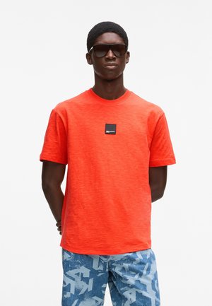 Camiseta roja de manga corta con cuello redondo y un parche de logo negro en el pecho. Combinada con pantalones de color azul claro con estampado.